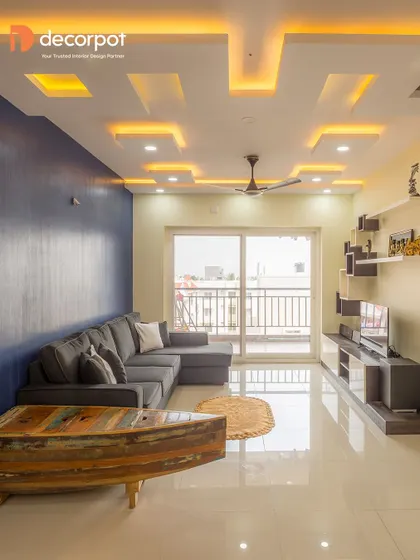 This living room design showcases a unique TV unit integrated with a geometric false ceiling. The blue accent wall adds a bold splash of color, while the boat-shaped coffee table is a quirky, conversational piece.