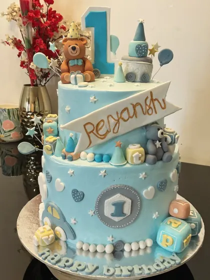 This two-tier first birthday cake is filled with cute details. It features a teddy bear topper, a toy train, building blocks, and a car, all in a lovely shade of blue.