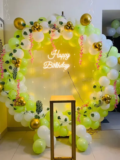 A fresh and vibrant lime green and white balloon ring. This setup is decorated with small floral and leaf details, a neon sign, and a pedestal for the cake.