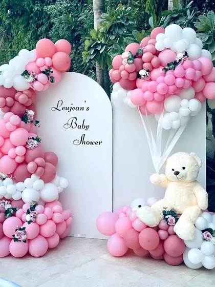 A stunning poolside baby shower. This elegant setup features a custom white backdrop and an elaborate pink and white organic balloon arch with a teddy bear accent.