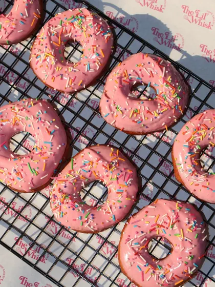 A whole tray of our freshly glazed pink donuts cooling on a wire rack, ready to be packed.