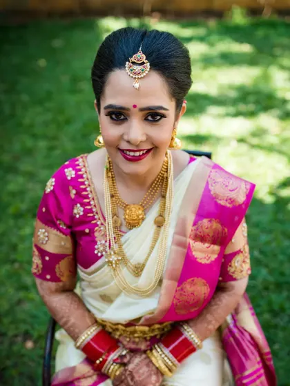 A delightful high-angle portrait of a smiling bride. Her traditional white and pink saree, layered gold necklaces, and happy expression make for a perfect wedding day memory.