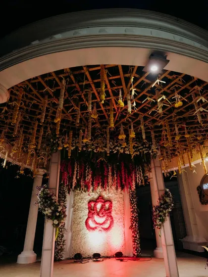 An elegant entrance to a wedding hall, featuring a floral Ganesha set against a wall of white chrysanthemums. The arched structure is decorated with hanging floral arrangements, symbolizing the sacred pillars of a mandap.