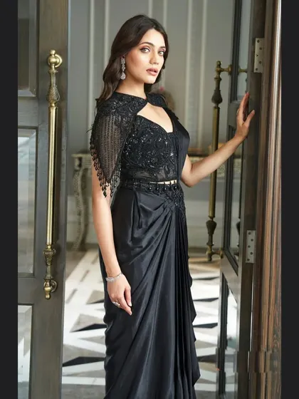 For a chic and modern reception or cocktail look, this black pre-draped saree is a perfect choice. It combines the elegance of a saree with the ease of a gown, featuring a stunningly embellished blouse with a stylish cutout and cape sleeves.