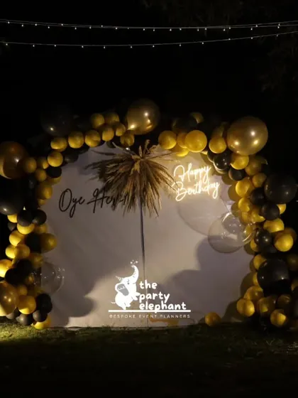 A centered shot of the black and gold photo backdrop, highlighting the symmetry of the design with its balloon arch and flanking floral arrangements.