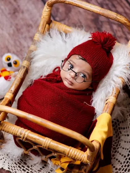 A creative and whimsical newborn portrait featuring a baby dressed in a Harry Potter theme. The baby is swaddled in red, wearing iconic glasses, and resting in a miniature bed surrounded by tiny books and an owl prop.