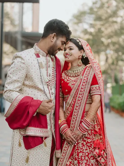A romantic couple portrait showcasing the bride's complete look. Her makeup is soft and timeless, a perfect match for her classic red wedding lehenga.