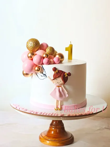 A sweet and simple first birthday cake featuring a cute fondant girl holding a cluster of pink and gold chocolate balloons.