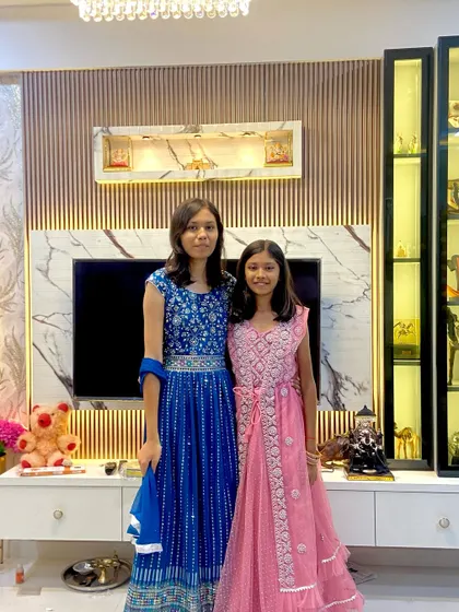 The Suryavanshi family's daughters enjoying their newly designed living room. The TV unit behind them features vertical fluting, marble accents, and a glass display case.