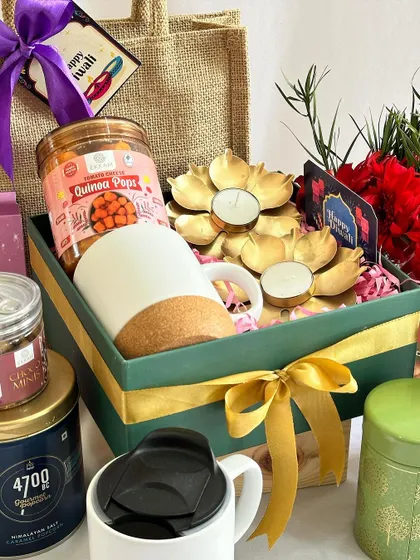 A festive gift box in green and gold, featuring a cork-base mug, quinoa snacks, and lotus-shaped diyas.