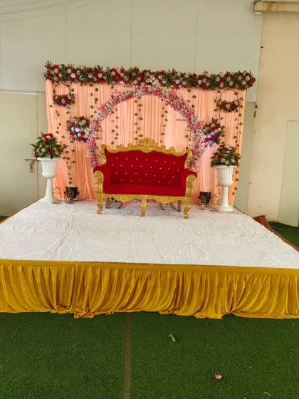 An outdoor wedding stage setup on an artificial grass lawn. The peach drapes and floral arch provide a soft, romantic backdrop against the open-air setting.