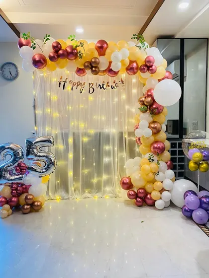 A beautiful 25th birthday decoration at home. The setup includes a balloon arch in peach, white, and metallic tones over a fairy light curtain.