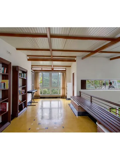 An upper-level corridor in 'Malenadu Mane' that doubles as a library and music space. The long horizontal window frames the landscape like a painting, and the exposed beam ceiling adds to the rustic charm.