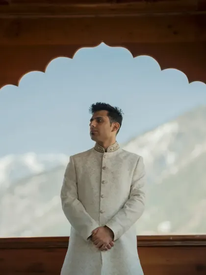 A handsome portrait of the groom in his ivory sherwani, framed by a traditional wooden archway against the mountain view.