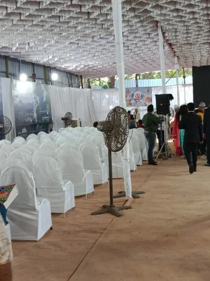 This image shows a wide view of a corporate event in progress. You can see the neat rows of fully covered chairs, pedestal fans for cooling, and fabric draped walls that create a professional environment inside a basic hall.