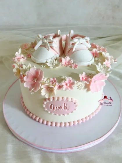 A cute baptism cake for a baby girl, featuring delicate pink sugar flowers and a pair of tiny, handcrafted baby shoes.