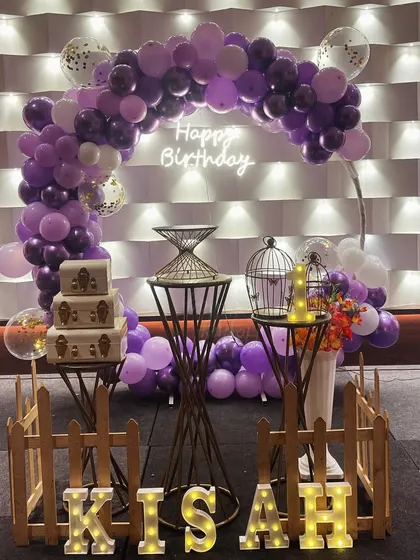 An elegant first birthday setup in purple and white, with a balloon arch, decorative props like birdcages, and marquee letters for the name.