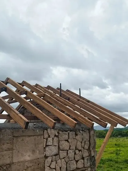 A building takes shape in harmony with its surroundings. Here, walls made of natural stone and rammed earth rise from the ground, while the wooden framework for the roof is put in place against a cloudy sky.