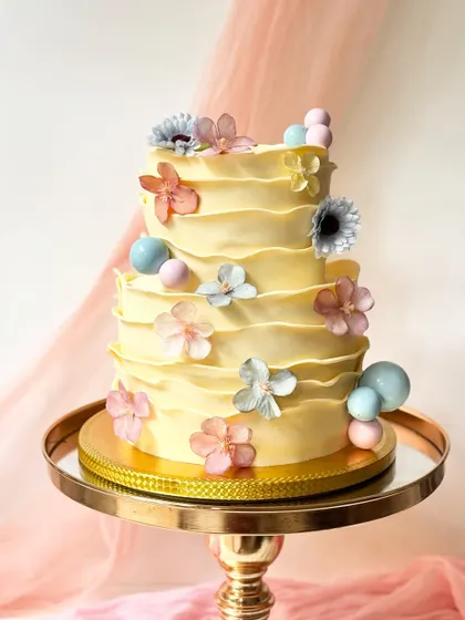 A cheerful three-tier cake with yellow ruffled buttercream. It's adorned with pastel-colored flowers and spheres for a playful, summery feel.