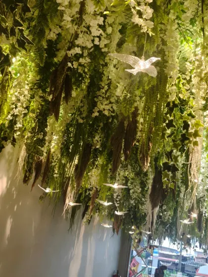 An enchanting ceiling installation for a lobby or entrance area. The dense hanging greenery and white flowers, interspersed with delicate glowing birds, creates a magical, forest-like atmosphere.