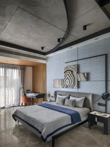 This well-lit bedroom in Swatantra Residence features a rhythm of blue tones against a raw concrete background. Dynamic artwork and rustic wood enhance the minimalist space.