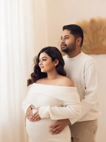 Looking toward the future together. The couple stands by a window, gazing outside, their hands joined over the baby bump in a pose full of hope and anticipation.