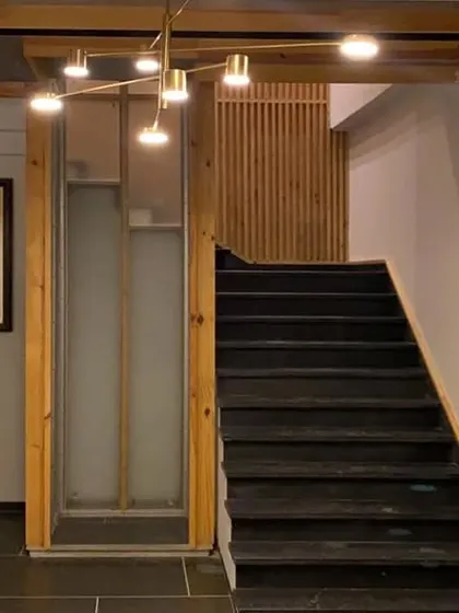 The staircase and elevator area in a Lucknow penthouse, framed by natural wood and a modern brass light fixture. A slatted wood screen provides texture and visual interest, demonstrating how fixtures can elevate transitional spaces.