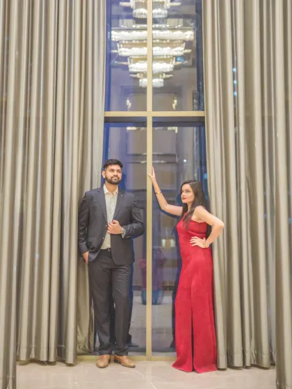 A dramatic, full-length portrait of the couple standing against floor-to-ceiling curtains. The window behind them reflects the city lights, adding a touch of urban glamour.