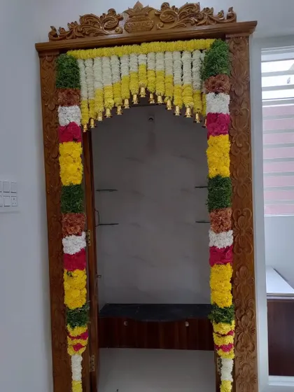 A full view of a pooja room door decorated with thick, colorful flower garlands. The bells hanging from the top add a traditional element.