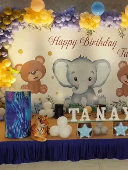 A cute and classic animal-themed first birthday stage. This setup features an elephant and teddy bear backdrop, a colorful balloon arch, and marquee letters spelling out the birthday boy's name.