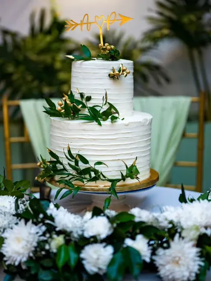 A simple and elegant two-tiered wedding cake, decorated with fresh greenery and a custom arrow-and-heart topper.