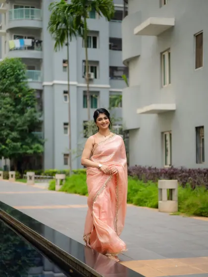 A full-length shot of actress Aditi Prabhudeva in a simple, elegant pink saree. The modern architectural background provides a clean and contemporary feel.