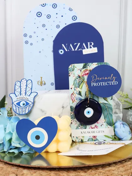 The complete Evil Eye Rakhi hamper, with a "Nazar" themed backdrop, a Hamsa hand sticker, and other protective charms.