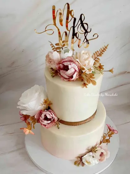 This two-tier wedding cake is a classic beauty, finished in smooth buttercream with a simple gold ribbon. The arrangement of fresh flowers adds a touch of timeless romance for your big day.