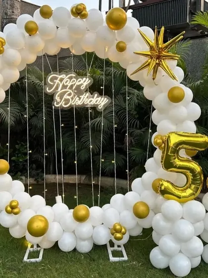 An elegant 53rd birthday decoration in a garden, featuring a circular white balloon arch with gold star accents and a 'Happy Birthday' neon sign.