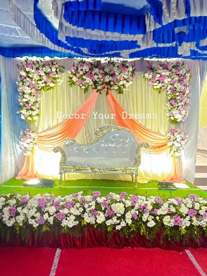 A beautiful outdoor engagement setup. The stage features a white and peach draped backdrop with floral bouquets, a classic chaise lounge, and a lush green carpet.