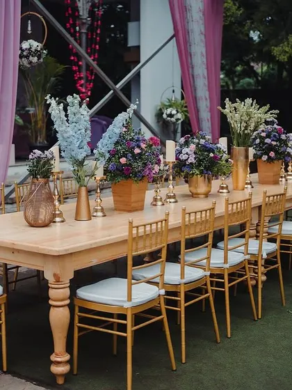 A long wooden table set for a reception dinner, with beautiful floral centerpieces and golden chairs.