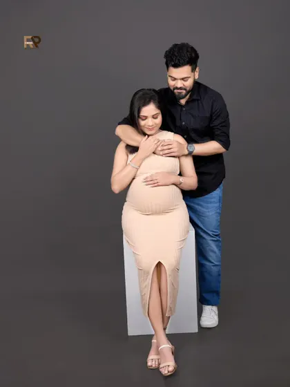 A tender moment between partners. The dad-to-be gently embraces his partner from behind, both focused on the baby bump. The neutral-toned outfits create a soft, warm feel.
