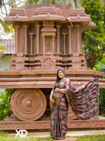 A dynamic shot of the mother-to-be with her saree flowing in the breeze, standing proudly before a traditional stone chariot.