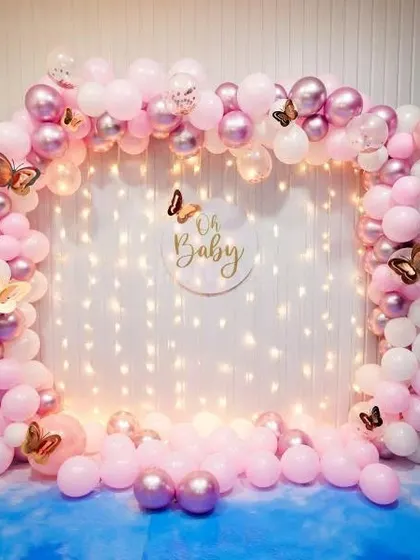 A delicate and pretty "Oh Baby" backdrop for a baby shower. The pink and white balloon arch is decorated with butterfly accents and set against a wall of fairy lights.