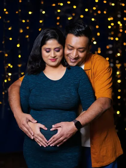 A cozy and romantic couple's portrait against a backdrop of warm fairy lights. The father-to-be embraces his partner from behind as they both create a heart shape with their hands on the baby bump.