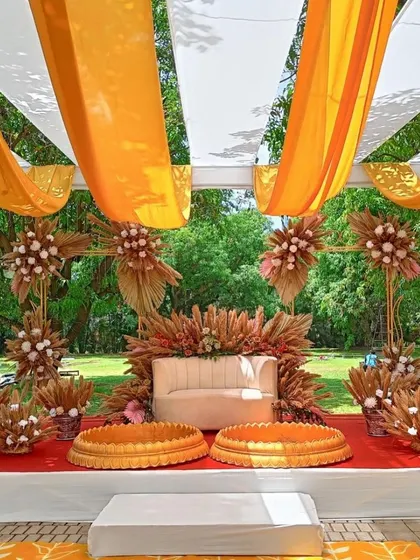 A full view of the boho-themed Haldi stage, showcasing the arrangement of pampas grass, a modern beige sofa, and traditional 'urli' bowls under a canopy of white and yellow drapes.