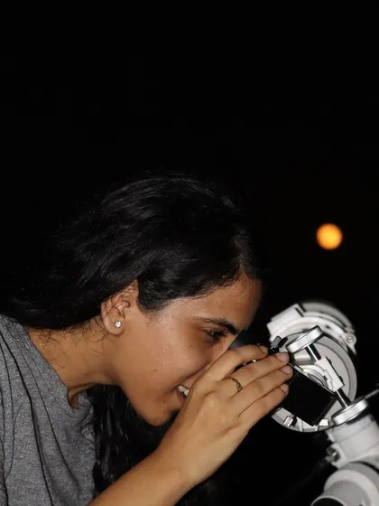 A woman looking through the telescope with the moon visible in the night sky behind her.