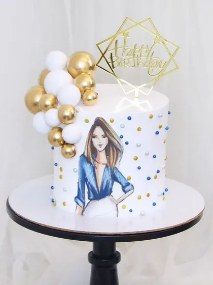 A chic and modern birthday cake for a stylish individual. The design features an edible print of a fashion illustration, complemented by a cascade of white and gold spheres and shimmering sprinkles.