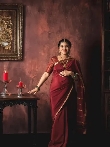 A solo portrait of a mother-to-be in a deep red saree, standing in a gracefully lit, vintage-themed studio setting that resembles a classic library.