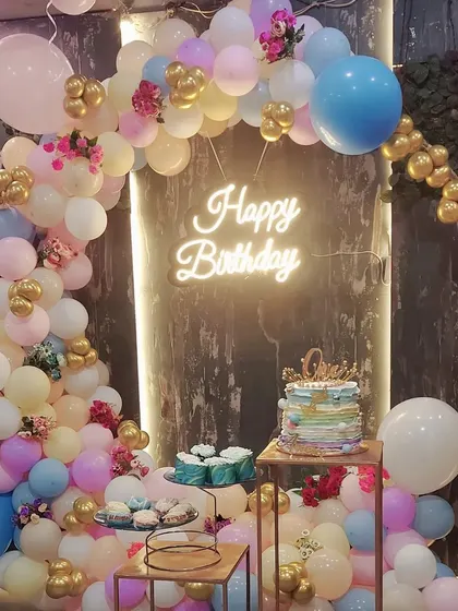 A complete party setup in a restaurant, featuring a pastel and gold balloon arch. The decoration includes a cake and dessert display, creating a perfect corner for birthday celebrations.