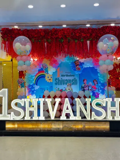 A grand Cocomelon first birthday celebration with large light-up marquee letters. The stage features a backdrop with a red floral overhang, creating a rich and festive atmosphere.