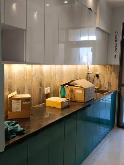 This kitchen, part of a 4BHK project in CGEWHO Kendriya Vihar, Greater Noida, is shown during the final stages of installation. It features a beautiful combination of teal green base cabinets and glossy white upper cabinets, complemented by a wood-textured tile backsplash.