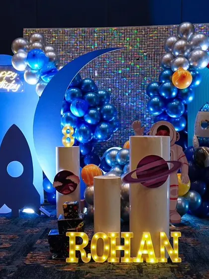 A cosmic adventure for Rohan's birthday with this outer space theme. The setup features a shimmering sequin backdrop, rocket ships, planets, and an astronaut cutout, all in a cool blue and silver color scheme.