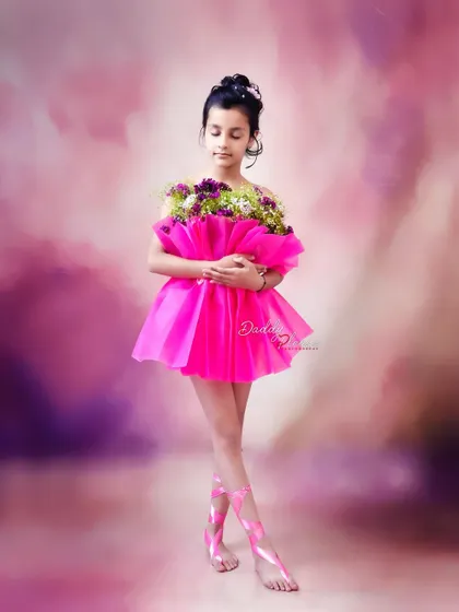 A full length shot showcasing the unique pink floral dress and matching ballet-style ribbons on her feet. The dreamy, cloud like background adds to the magical feel.